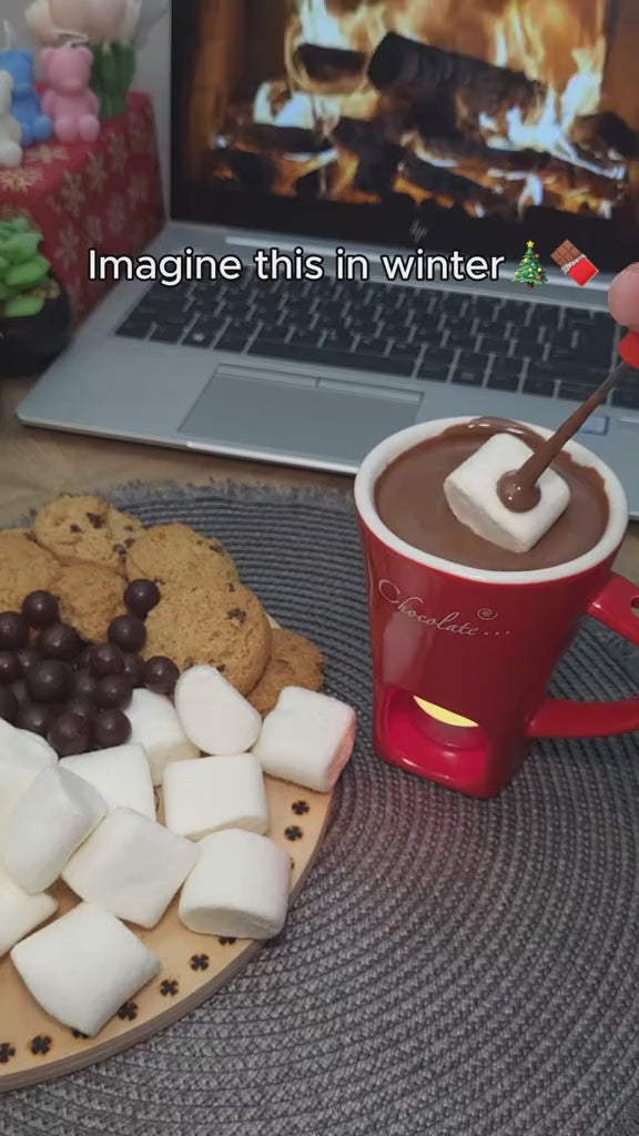 Chocolate melting in the red fondue mug over a tealight, with cookies and marshmallows, in front of a fireplace video